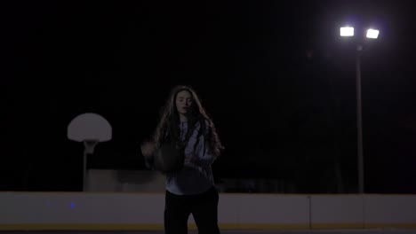 teenage girl with long brown hair dribbles ball, throws it up and catches it before shooting at a dark outdoor court with lights