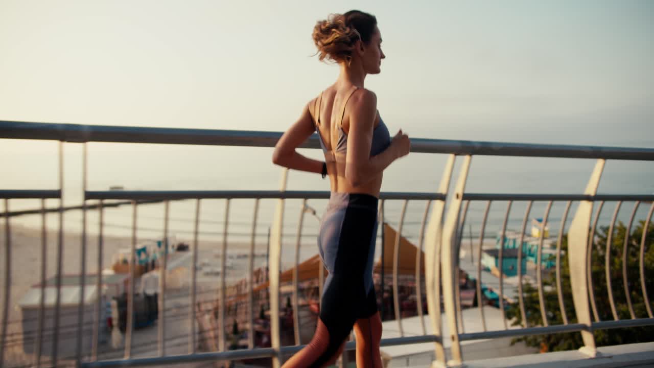 스포츠 유니폼을 입은 소녀가 해변에서 격자 울타리를 따라 달린다. 해변에서 아침 달리기