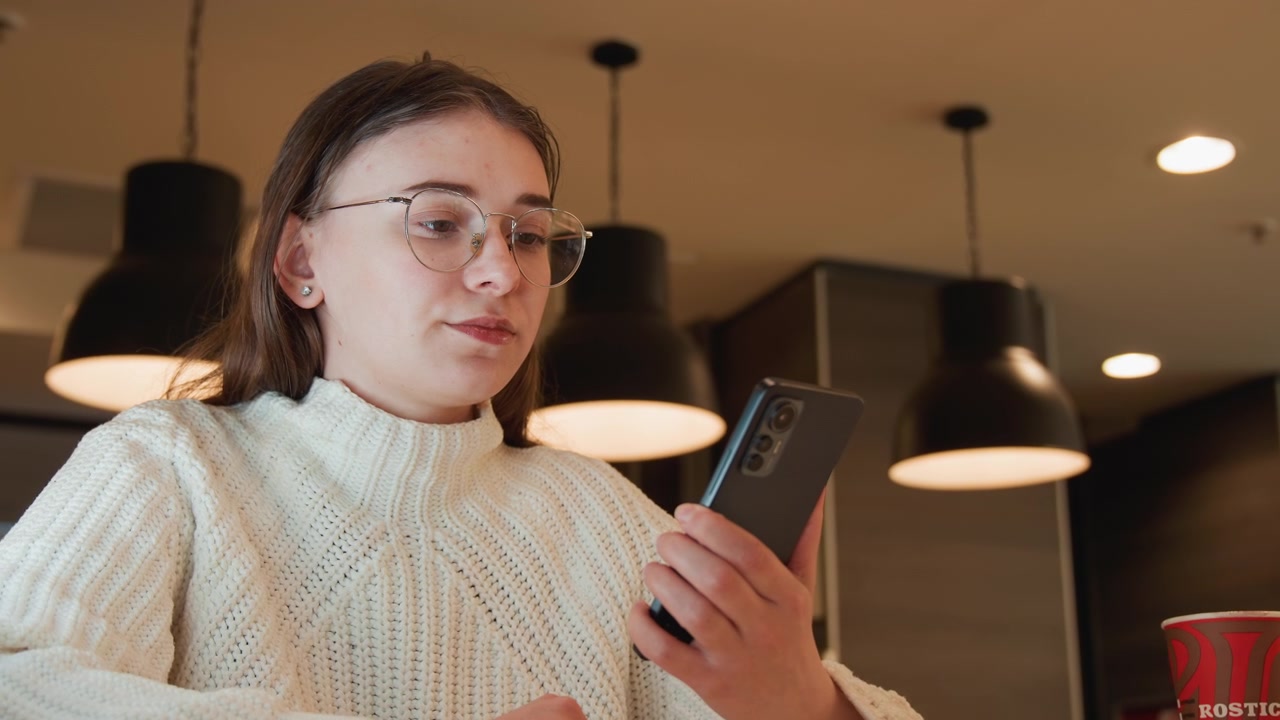 Jovem mulher com suéter de tricô grosso e óculos senta-se à mesa de madeira em um interior de cozinha aconchegante, estendendo a mão em direção ao smartphone enquanto olha para baixo com expressão calma e focada e postura relaxada
