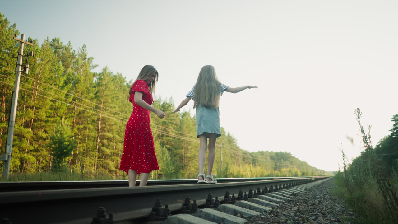 back view mum in red dress watching daughter balancing with arms outstretched as she walks carefully on railway beam flanked by lush green forest under soft sunlight for playful family bond