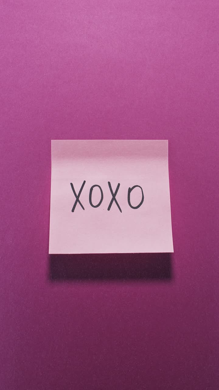 Hands posting XOXO sticky note on bright pink wall, capturing affection concept