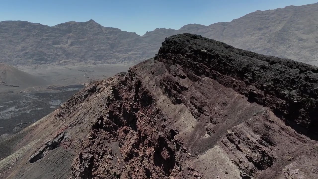 Ripresa con drone che vola molto vicino al bordo stretto e frastagliato del cratere del Pico do Fogo, mettendo in evidenza la cresta basaltica affilata e l'ampio paesaggio vulcanico di Capo Verde sullo sfondo.
