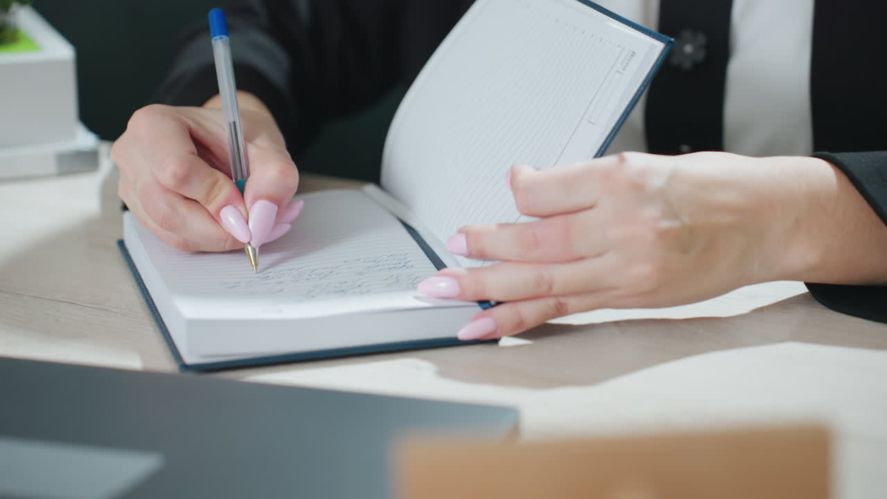 close up of adult woman with long pink nails taking down handwritten notes in lined journal while seated at desk with open laptop and architectural building model slightly visible in background