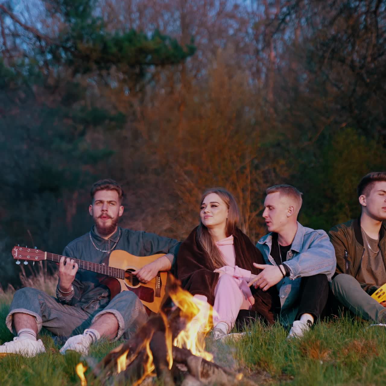 Serata accogliente di amici seduti accanto al fuoco. Giovani che cantano una canzone insieme mentre un uomo suona la chitarra su sfondo di alberi
