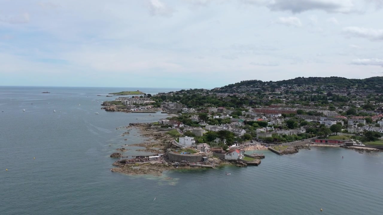 Vuelo aéreo amplio a lo largo de un suburbio costero de Dublín con una torre Martello, una cala rocosa para nadar y barcos en alta mar en un día gris y tranquilo