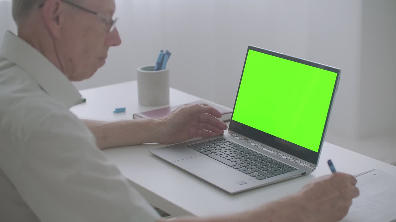 old man is looking at screen of laptop and writing notes in notebook, chroma key technology for post-production