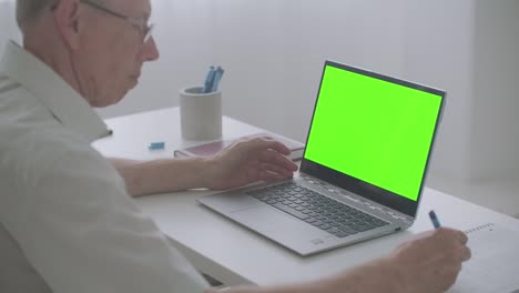 old man is looking at screen of laptop and writing notes in notebook, chroma key technology for post-production