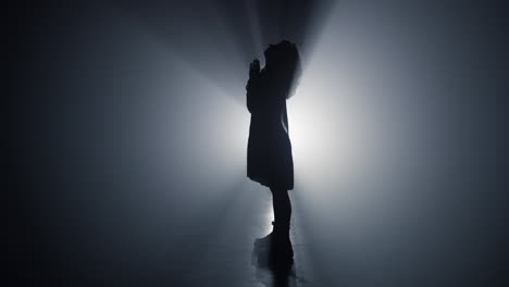 silhouette praying girl indoors. female person folding hands for prayer in dark.