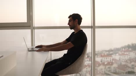 Relaxed-caucasian-man-in-stylish-glasses-using-laptop,-having-video-call.-Cheerful-smiling-man-in-black-clothes-sitting-on-chair-having-a-friendly-video-call.-Happy-man-enjoying-video-calling-at-big,-bright-house