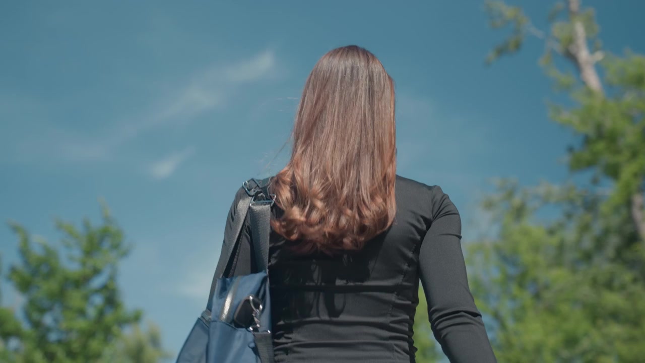 Close up of woman with long hair walking outdoors on sunny day carrying blue bag. Hair flows naturally in wind wearing black top and sunglasses. Background features clear sky trees creating
