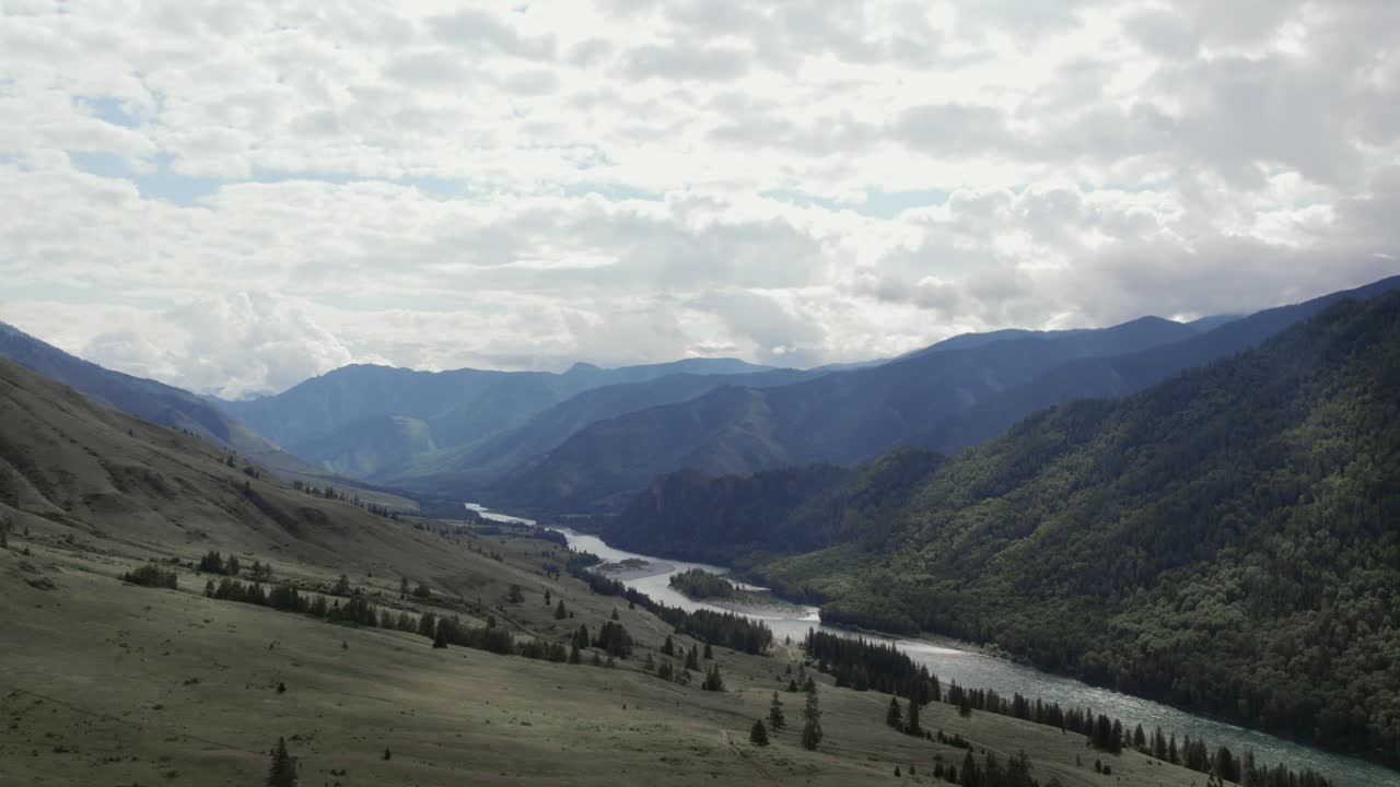 Wide aerial landscape of the Ak-Kem valley with a winding river, forested slopes, and dramatic clouds over the Altai Mountains in the Altai Republic, Russia