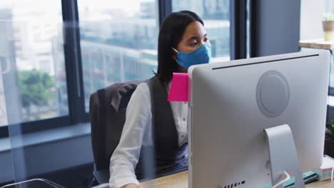 mujer con máscara facial usando una computadora mientras está sentada en su escritorio en una oficina moderna