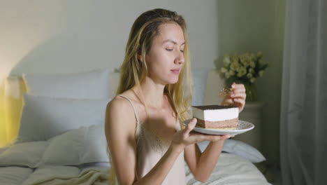 chica rubia sosteniendo y comiendo un trozo de pastel sentado en la cama