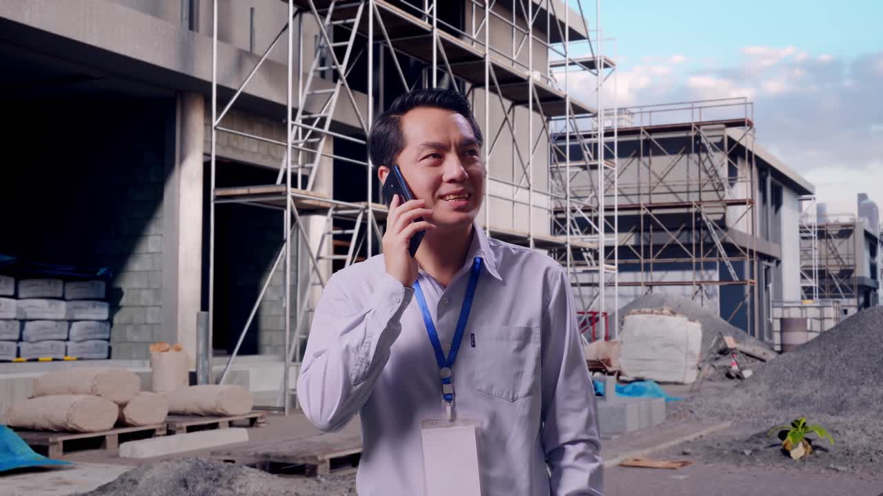 Trabajador profesional asiático de pie en una obra en construcción de una nueva casa; coge el teléfono y empieza a hablar.