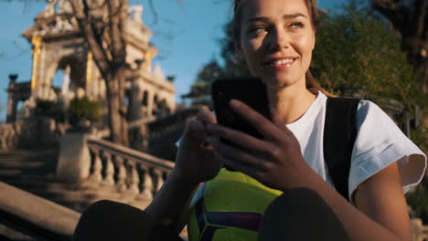 Porträtaufnahme-Einer-Hübschen-Frau-Mit-Fußball-Und-Rucksack,-Die-Nach-Dem-Training-An-Einem-Sonnigen-Tag-Ihr-Smartphone-Benutzt-Und-Wegschaut.