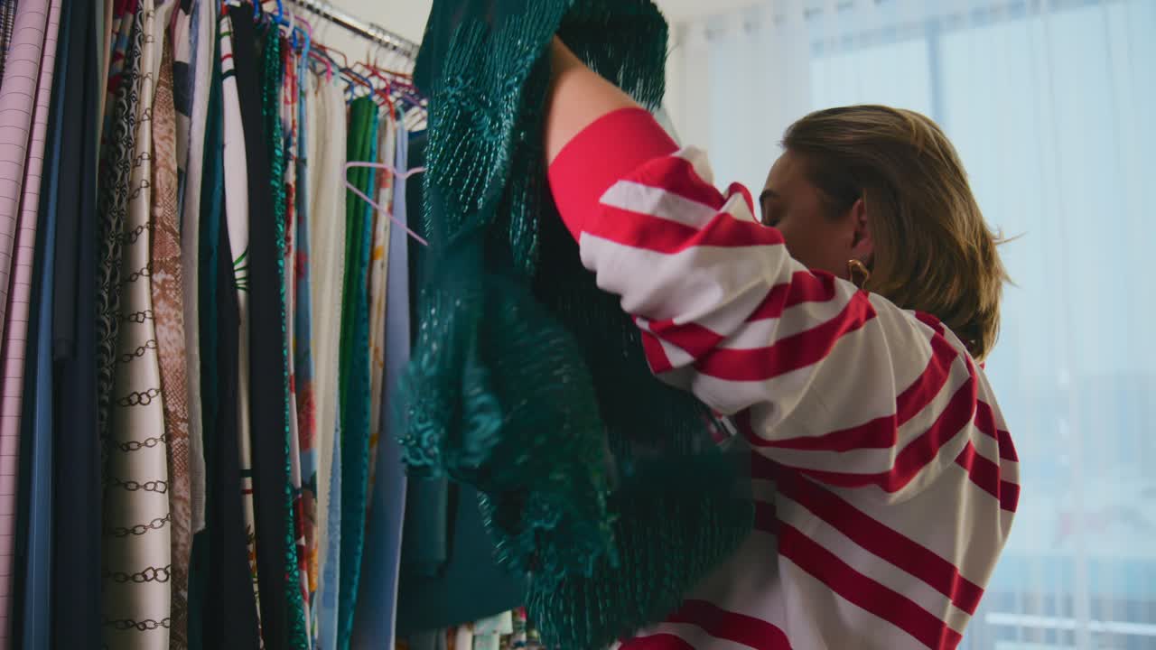 Woman browses fabrics on a rack, selecting a green piece. She examines textures, pausing to feel material quality. Bright window light fills room, enhancing vibrant colors. She seems focused, calm