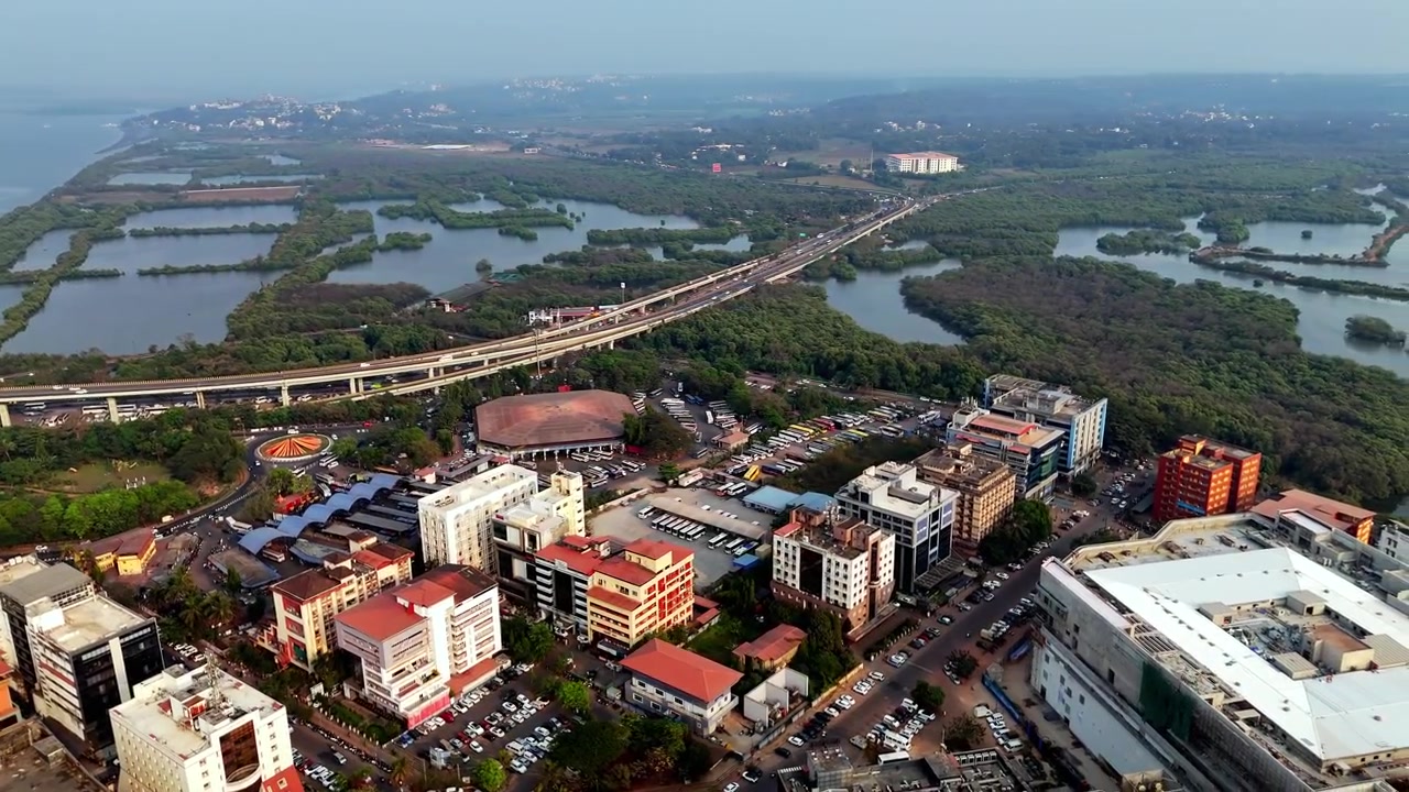 Veduta aerea cinematografica della stazione degli autobus KTC circondata da edifici moderni, strade trafficate e aree di parcheggio, con autostrade sopraelevate che si estendono verso zone umide e paesaggi lontani.