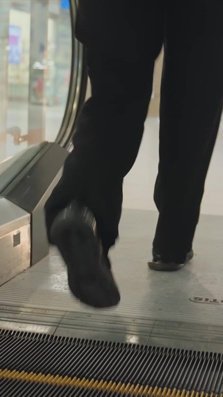 Person in dark pants and shoes stands on moving escalator. Subject progresses through space in dark shoes clasping. Tiled surface is light colored with indistinct objects