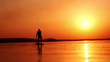 black male silhouette of a sportsman practicing sup boarding. man holding a paddle rowing with it. orange sun and sky at backdrop.