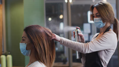 female hairdresser wearing face mask spraying hairspray on hair of female customer at hair salon