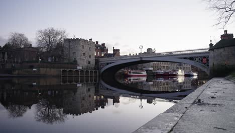 Low-push-in-towards-perfectly-still-River-in-historic-city-with-traffic-passing-over-bridge-at-sunrise