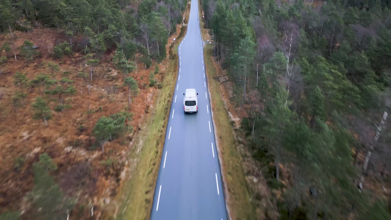 A camper van drives down an empty road through a forested autumn landscape, aerial view