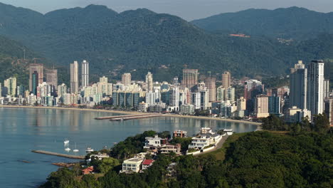 Aerial view capturing Meia Praia beach in Itapema, Santa Catarina, Brazil, with calm sea reflecting buildings, boats sailing near the shore, and mountains in the background