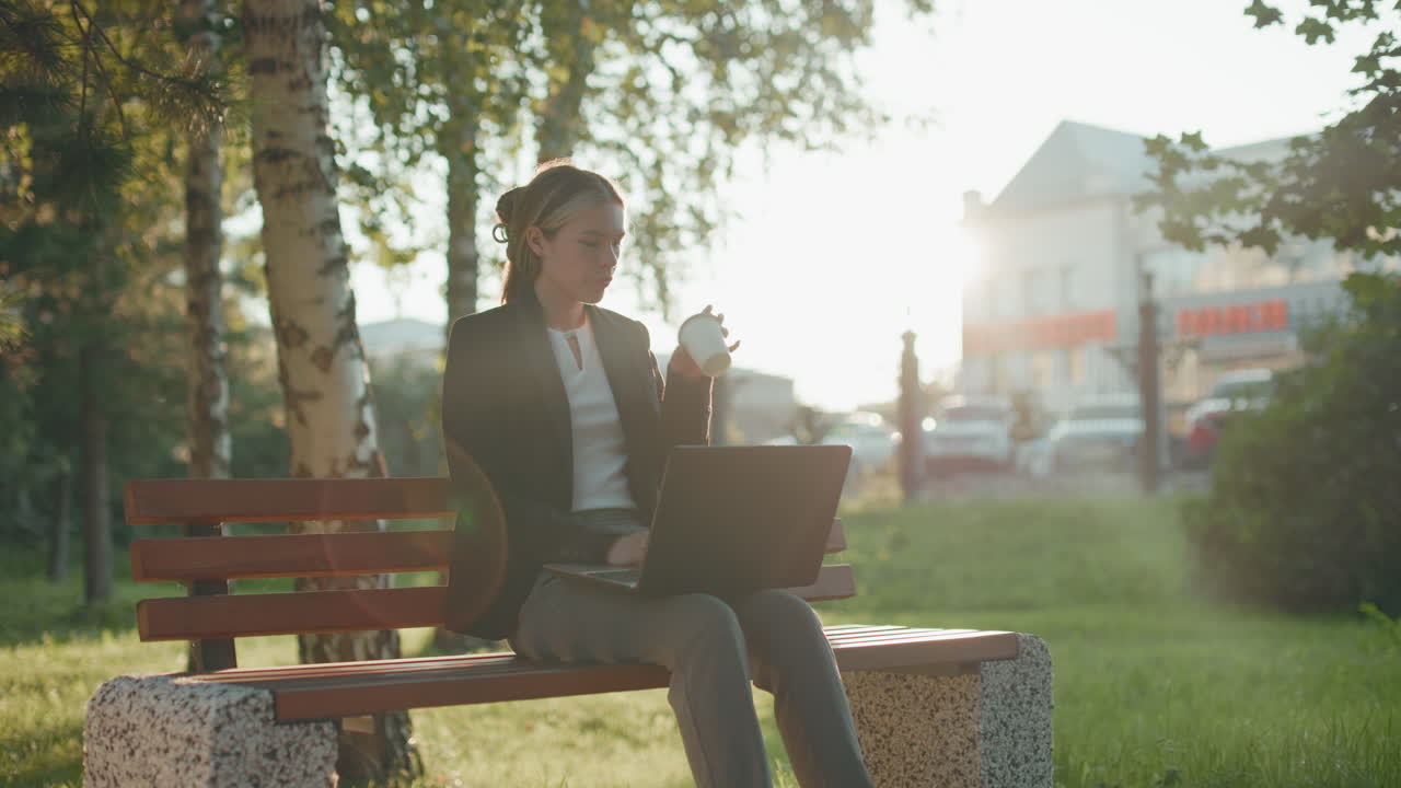 Silhouette effect surrounds woman working outdoors in park while sipping beverage from cup with laptop on lap and blurred background featuring buildings, trees, and cars