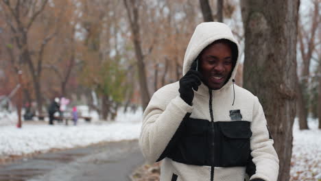 excited young man chatting over phone, chewing gum, with blurred background of trees, buildings, and people walking in urban setting, showcasing a joyful moment in daily life