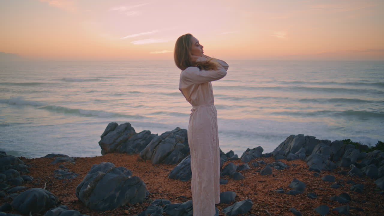 아름다운 여자가 바다 절벽의 저녁 풍경을 포즈하고 있습니다. 해가 지는 것을 즐기는 편안한 소녀