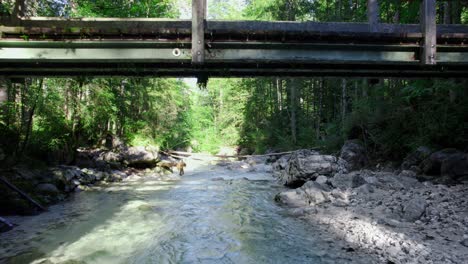 Aerial-drone-shot-going-under-a-small-wooden-bridge-over-a-flowing-stream-through-a-lush-green-forest-at-daytime