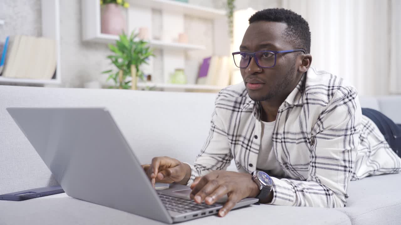 Handsome African man sitting on sofa in living room at home working with laptop.