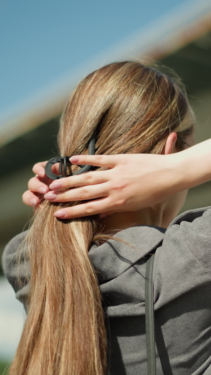 회색 셔츠를 입은 여인이 다리 근처에서 머리카락을 모으기 위해 헤어 클립을 사용하는 측면 뷰, 배경은 다리의 구조와 함께 약간 흐릿합니다.