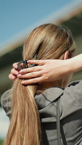 vista lateral de una mujer con una camisa gris mientras usa un clip para el cabello para recoger su cabello cerca de un puente, el fondo está ligeramente borroso, con la estructura del puente
