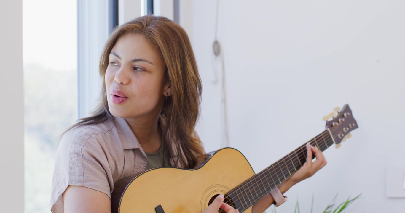 Donna afroamericana che suona la chitarra acustica, cantando vicino a una finestra alta, reagendo a una voce fuori campo.