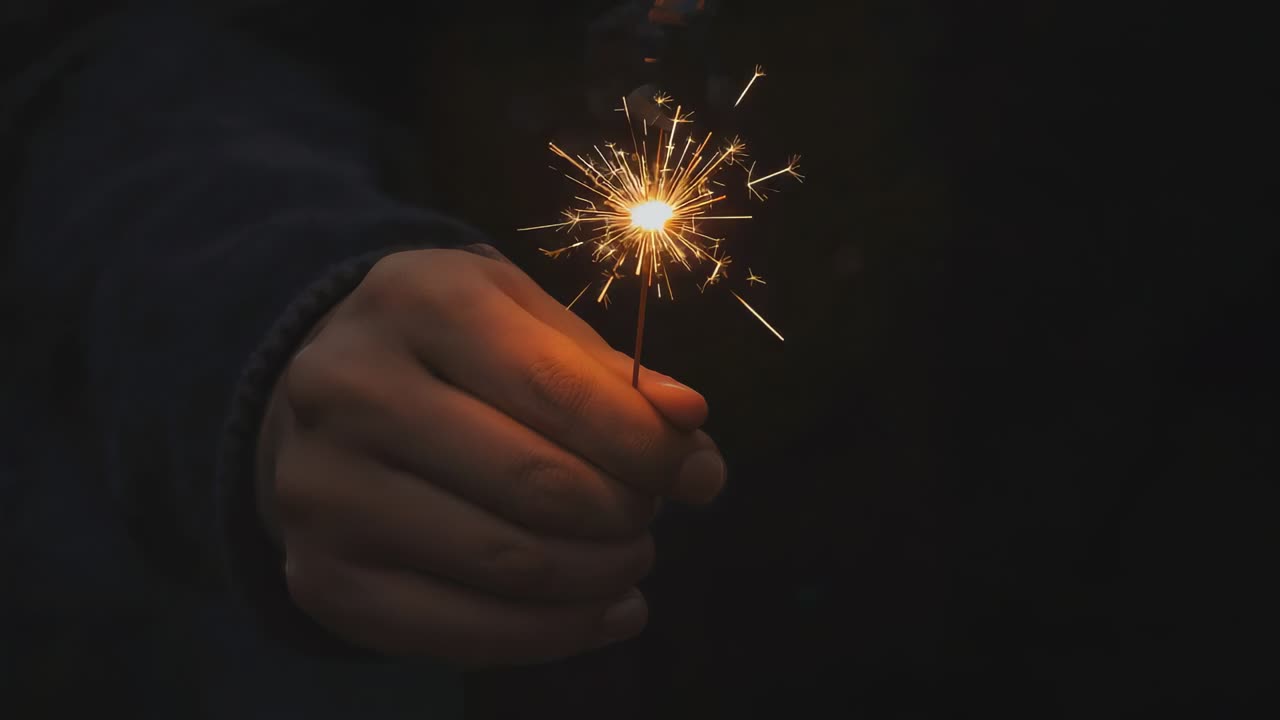 Holding hand in dark long-sleeve, gripping sparkler igniting, sparks detaching at night