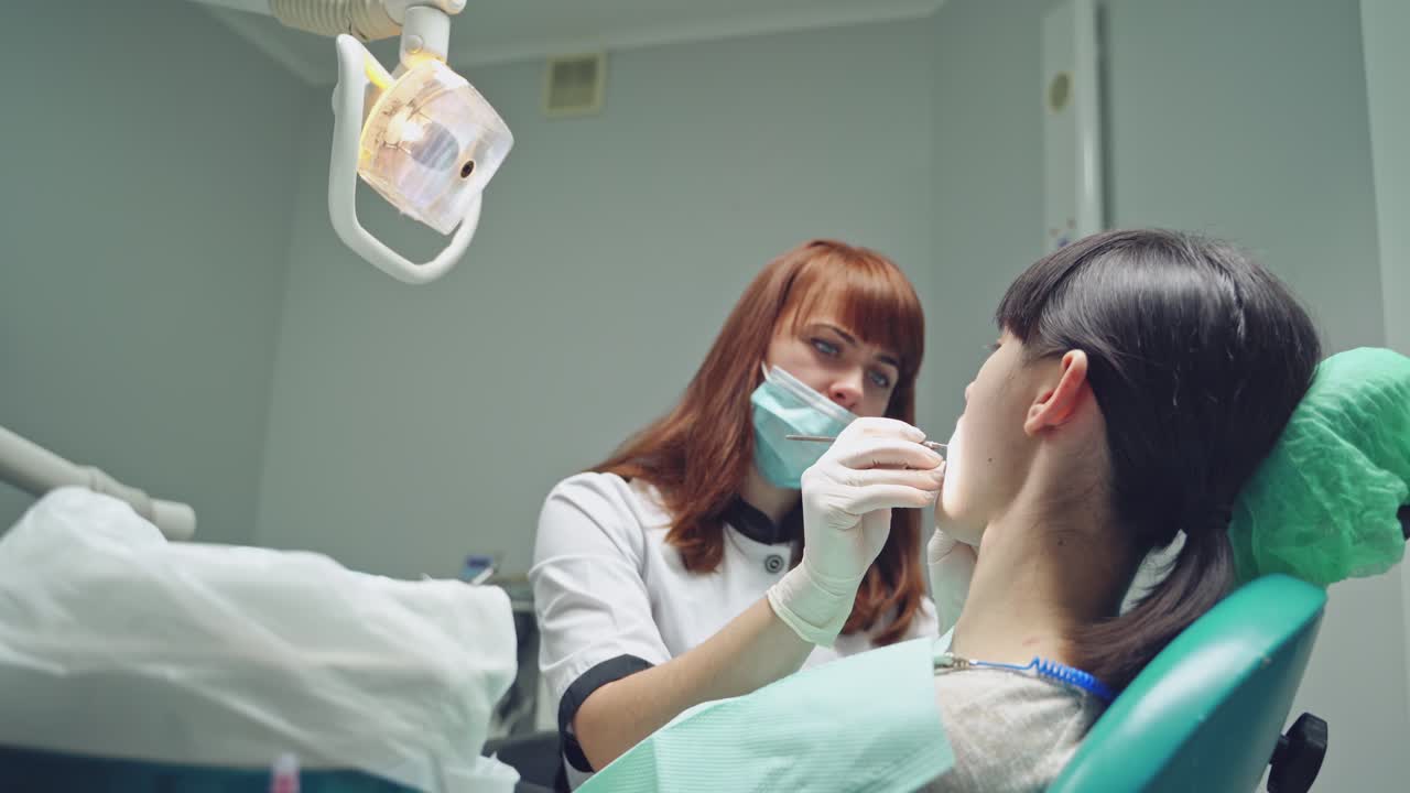 Red-haired female dentist looking at patient's teeth before treating them in stomatology office. Dentist treating teeth of woman patient.