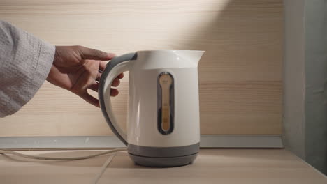 close-up of man in bathrobe holding electric kettle with water level indicator. minimalist kitchen setting with soft lighting, focusing on clean design of kettle and kitchen countertop