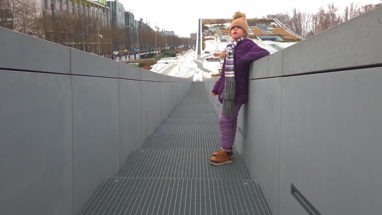 En ung jente løper selvsikkert langs en betongsti i en snødekt urban park om vinteren. Kaldt vær, snødekt landskap og bybakgrunn skaper en aktiv, leken sesongscene