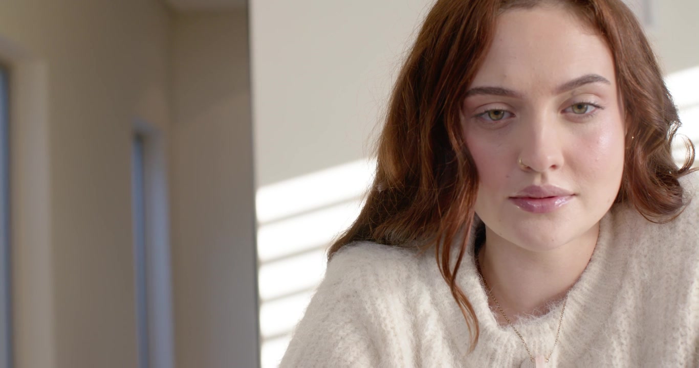 Woman sitting by window in room sun bands cream sweater tilting gaze at laptop during recording
