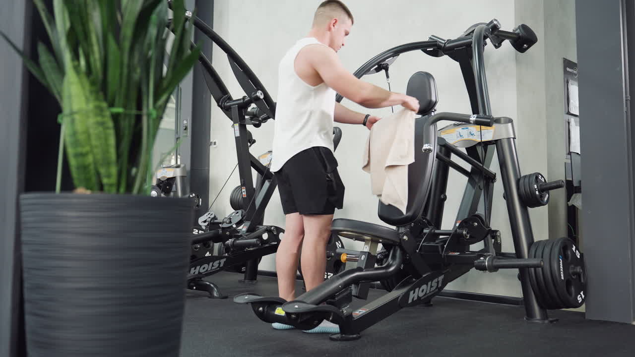 Gym goer walks toward machine and spreads towel on seat next flower vase in corner while preparing for intense workout session amid modern fitness equipment with sleek metal frames