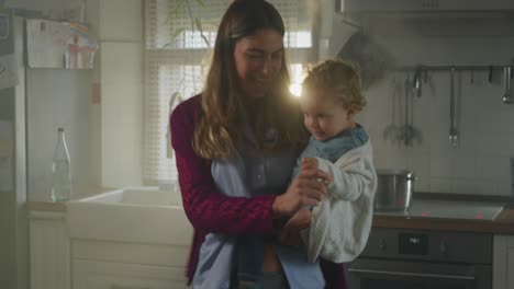 cinematic shot of an young happy smiling mother is keeping in arms her toddler baby boy and having fun to dance together in a kitchen at home. concept of  happiness, love, childhood, life, motherhood.