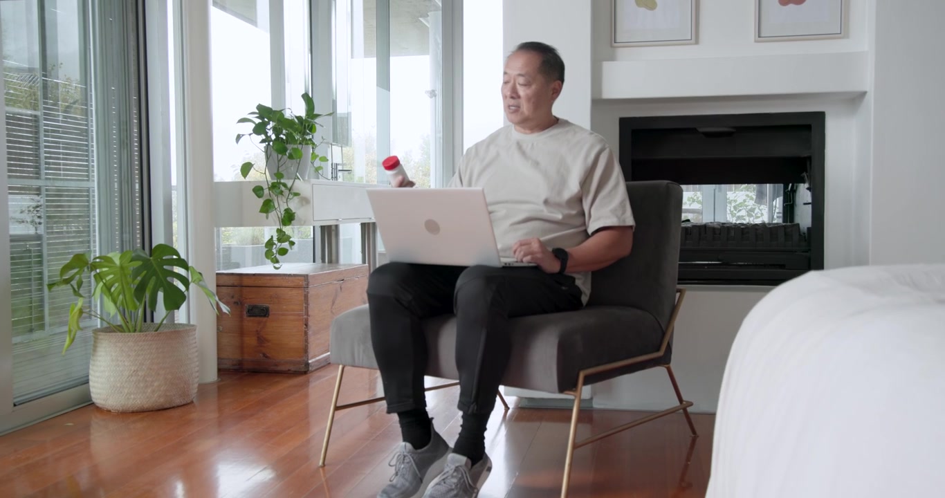 Senior Asian man sitting in bedroom armchair holding bottle with red cap checking laptop dosage