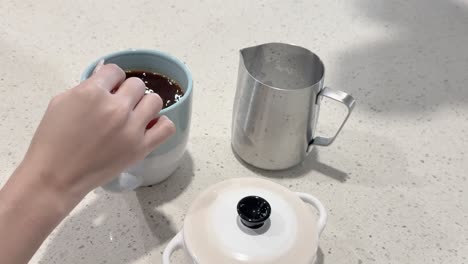 a hand stirs sugar into a coffee cup using a spoon on a kitchen counter