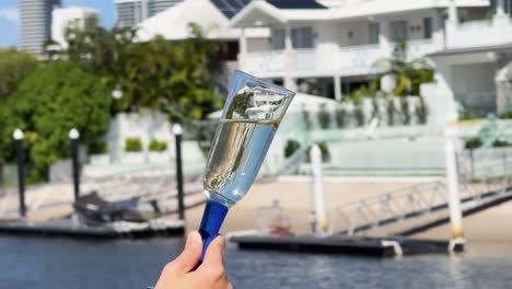 a hand raises a champagne glass filled with sparkling wine against a sunny waterfront mansion backdrop. bright daylight, smooth camera movement, celebratory mood