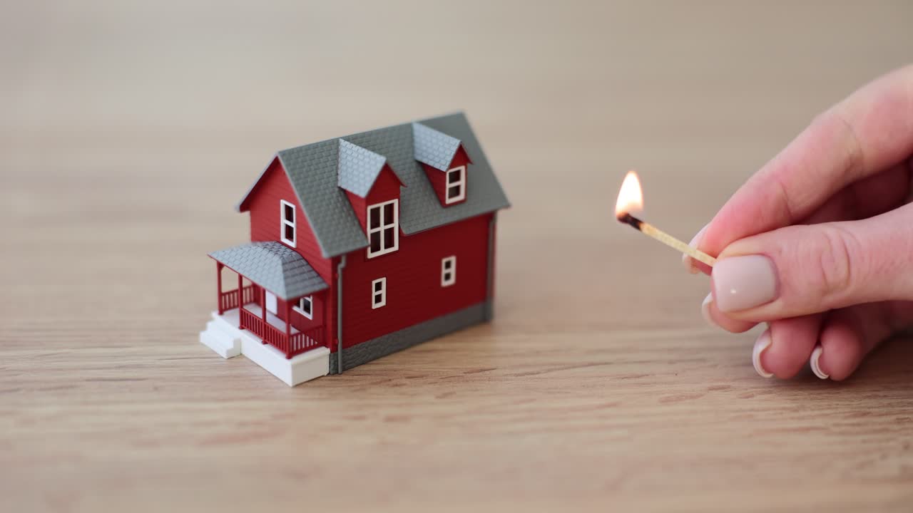 Une main de femme approche une allumette allumée d'une maquette de maison sur une table en bois