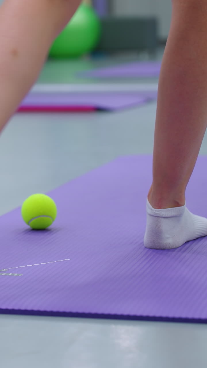 vista de la pierna trasera en calcetines moviéndose hacia adelante y hacia atrás en una alfombra púrpura con una pelota de tenis verde, el espejo del gimnasio refleja el fondo, capturando la sesión de fitness y el movimiento de los pies durante el ejercicio de coordinación