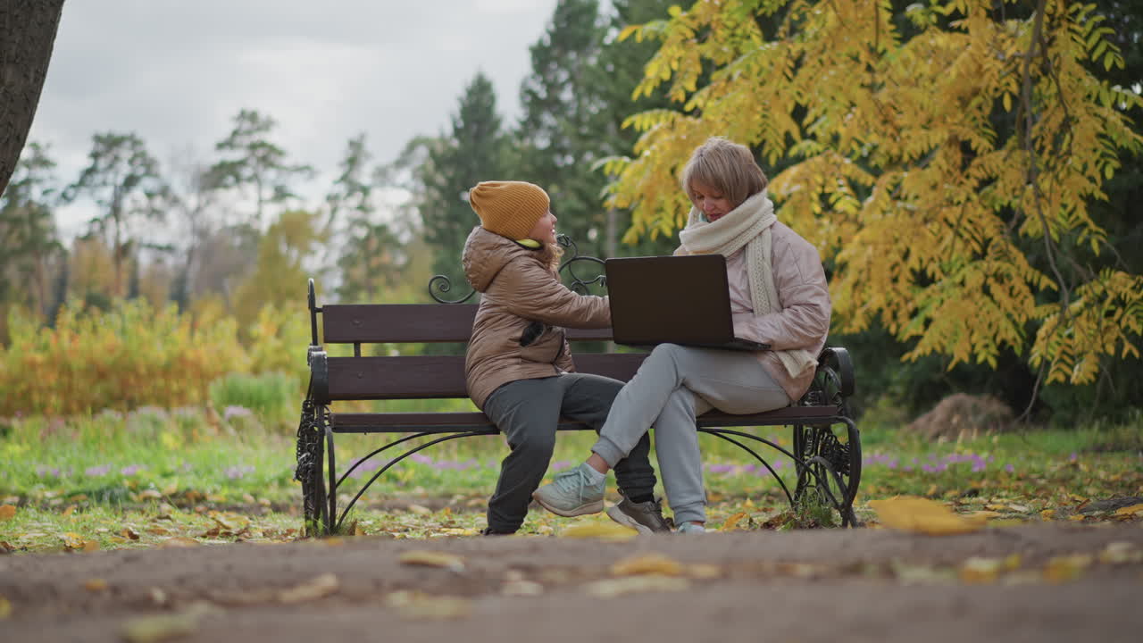 little girl running toward mum working on laptop on ornate bench in autumn park holding bright yellow leaf excitedly amid scattered foliage and golden trees