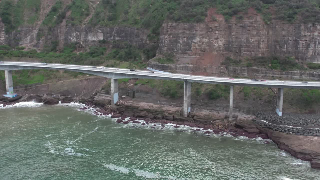 Aerial view of modern coastal bridge stretching over ocean, with lush green hills, rocky shore, and clear blue water, highlighting infrastructure and scenic natural landscape