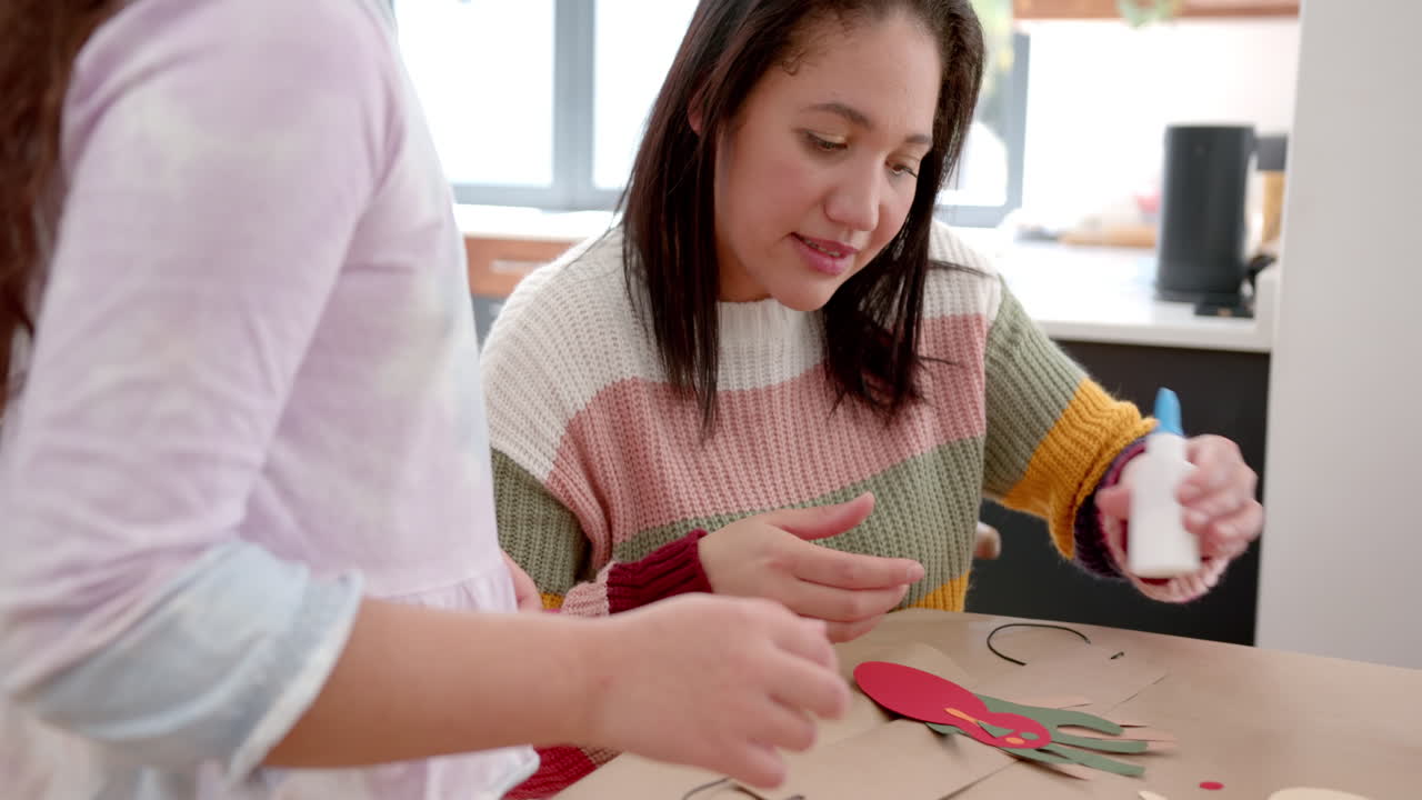 madre y hija bi-raciales felices pegando recortes coloridos en la sala de estar soleada